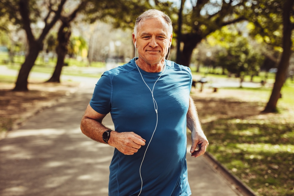Senior man running in a park, wearing a blue shirt and earphones, enjoying a healthy lifestyle.