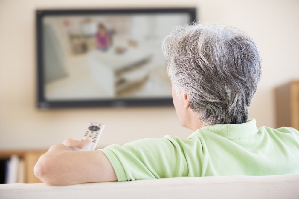 Senior man watching television, holding a remote control while sitting on a couch at home.