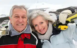 An elderly couple enjoying a day of skiing and winter outdoor exercise in a snowy mountainous landscape, dressed in winter gear and smiling.